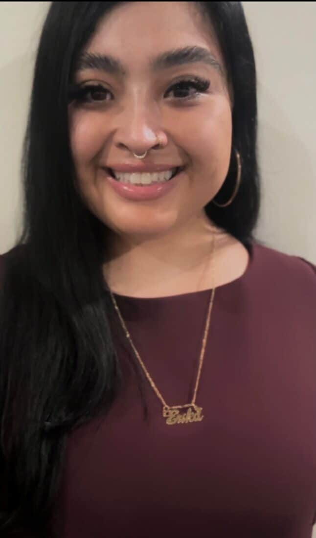 A woman with long dark hair is smiling at the camera. She is wearing a maroon top, large hoop earrings, and a gold necklace with a nameplate. She has a septum piercing and is standing against a plain light-colored background, representing The Representation Project's Board of Directors.