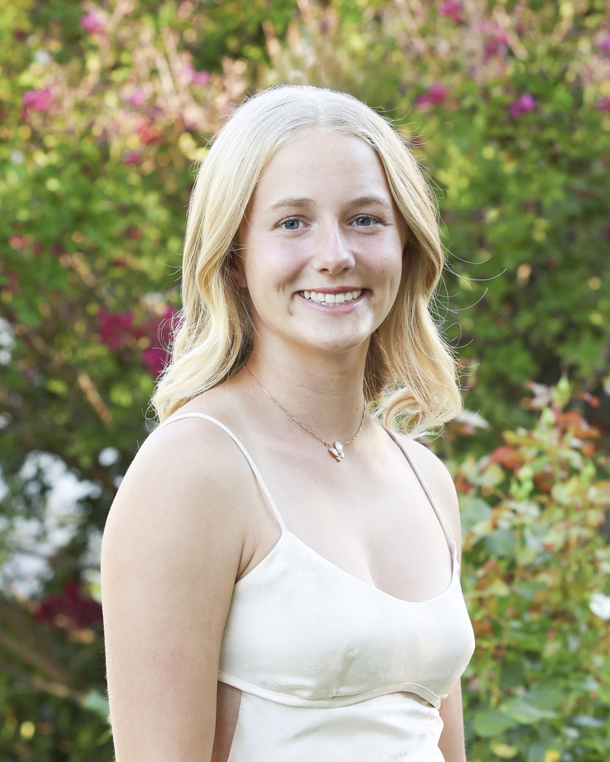 Kate Ragatz, a member of The Representation Project's Youth Advisory Council, smiles while standing outdoors amidst green foliage and pink flowers.