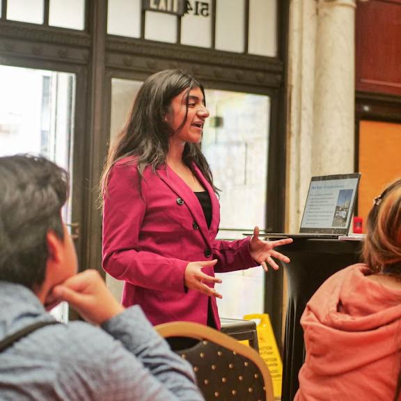 Maegha Ramanathan, a member of The Representation Project's Youth Advisory Council, gives a presentation in front of an audience as she stands next to a laptop on a podium.