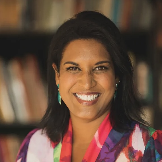 A woman with medium-length dark hair, wearing a colorful floral top and turquoise earrings, smiles brightly in front of blurred bookshelves—an inspiring member of The Representation Project's Board of Directors.