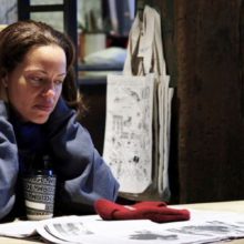 Still from "On the Record" movie. A woman sits at a table and looks at an open newspaper.