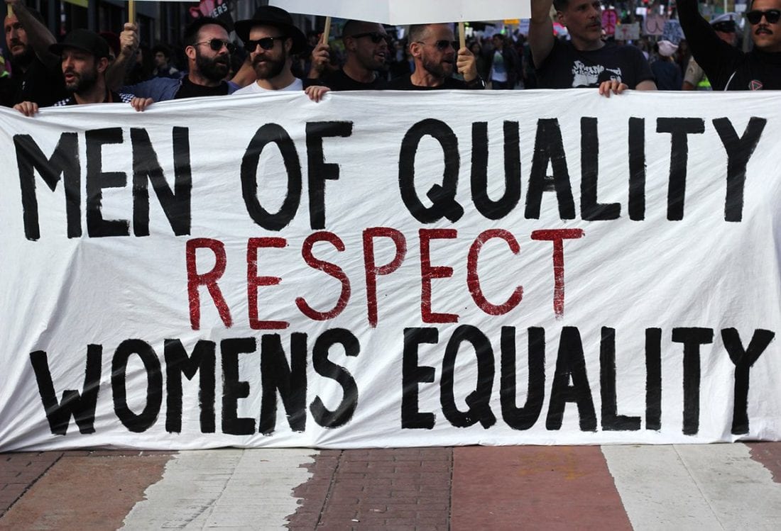 Men marching in allyship with women. Several men hold up a sign together: "Men of Quality Respect Women's Equality"