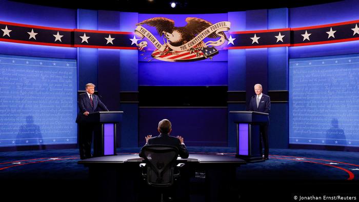 Still from presidential debate. Trump at the left podium, Biden at the right podium. A moderator is seated between them at the front of the stage.