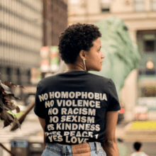 A person stands in an urban setting wearing a black t-shirt with white text that reads "No Homophobia, No Violence, No Racism, No Sexism, Yes Kindness, Yes Peace, Yes Love." This embodiment of values and inclusivity is set against a backdrop of a street with tall buildings and a potted plant with flowers.