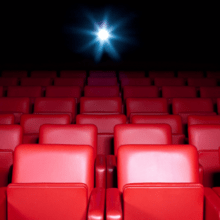 Empty red theater seats face a brightly lit screen in a dark cinema. The glow from the screen illuminates the rows, creating a contrast between the vibrant red seats and the dim surroundings, awaiting the keen eye of a female film critic to breathe life into its narrative.