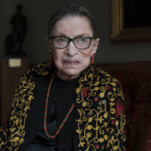 An elderly woman with short, pulled-back hair and glasses is sitting on a leather chair. She is wearing a black outfit with a decorative, embroidered shawl and red jewelry. The background features a dark statue and a framed portrait of RBG, portraying her as an inspiring role model for women everywhere.