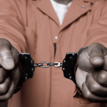 A black person in an orange jumpsuit holding out their wrists, which are handcuffed.