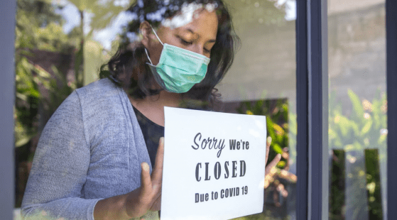 A picture of a woman in a face mask putting a sign in a store window. The sign says "Sorry, we're closed due to COVID-19"