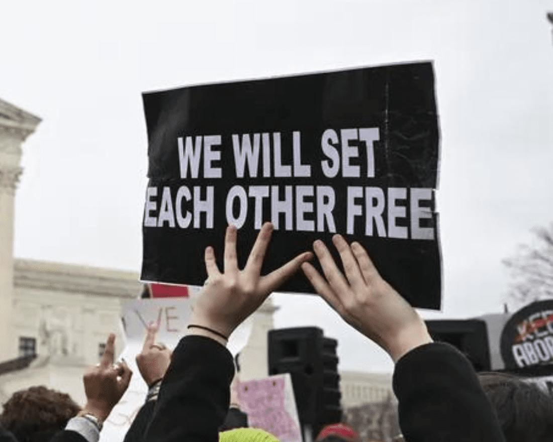 A person holds a sign up with both hands. The sign reads, "We will set each other free."