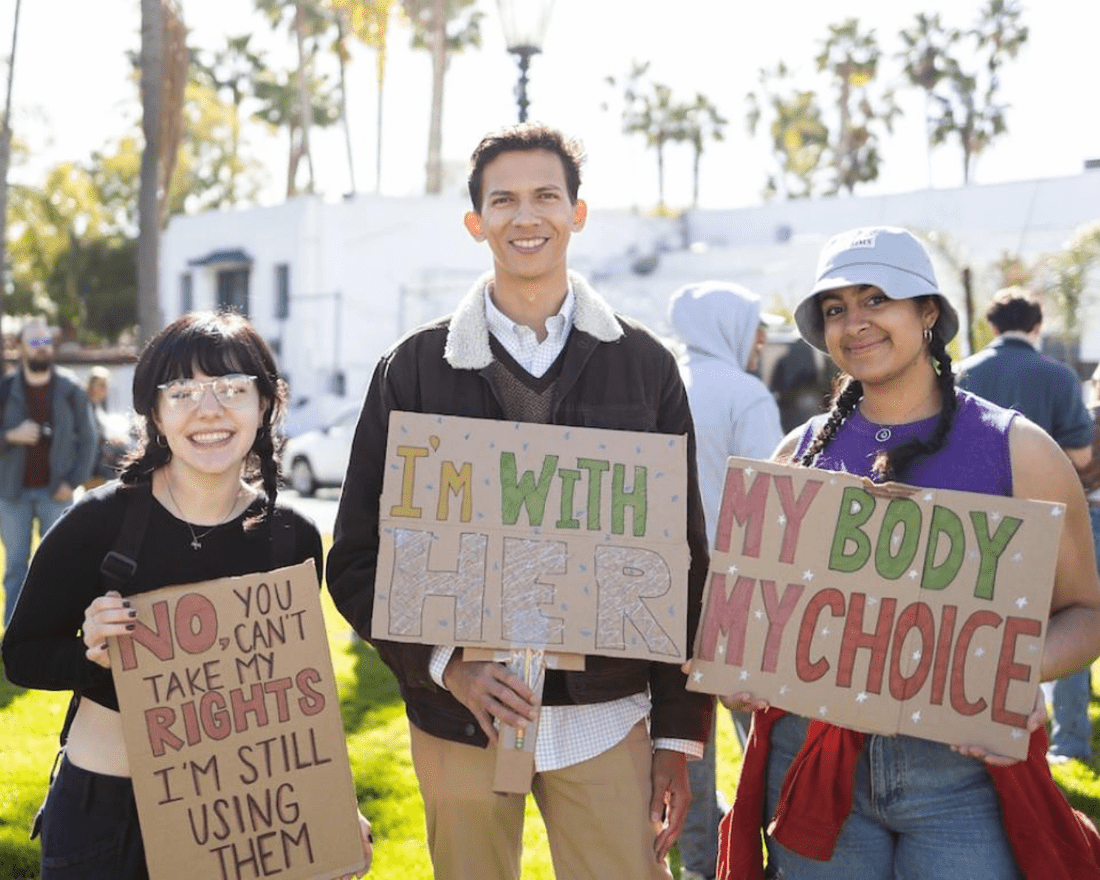 Three people stand side-by-side and pose with their signs. From left to right, the signs read: "No you can't take my rights, I'm still using them," "I'm with her," and "my body my choice."