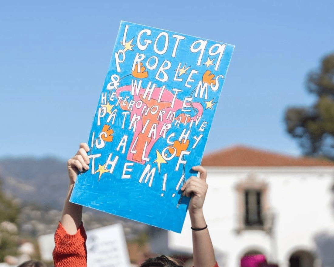 A person holds up a sign with both hands. The sign is blue with a uterus, stars, and flowers drawn on and text written on top. The text reads: "I got 99 problems and white heteronormative patriarchy is all of them!!"
