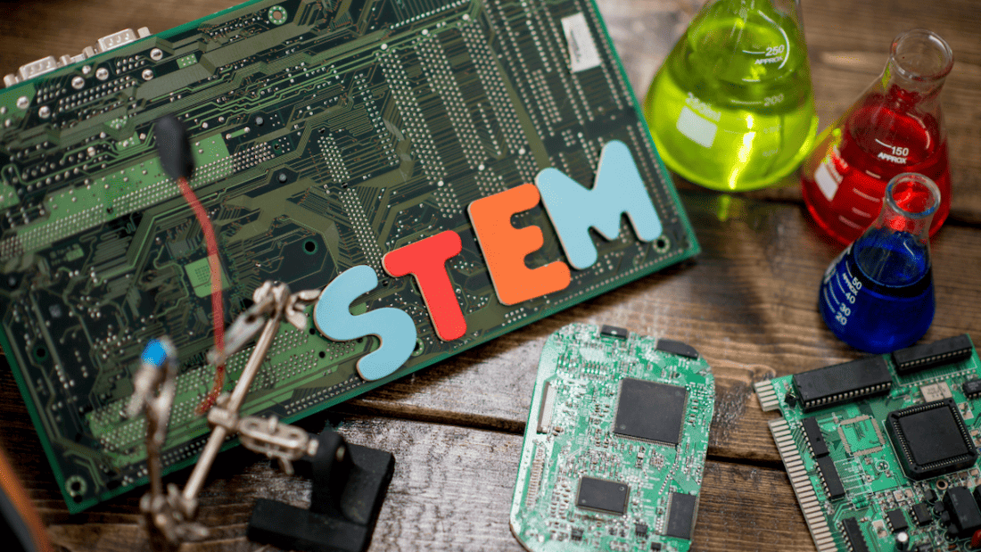 A variety of science-related materials are displayed on a table, including: a large motherboard, science flasks filled with color liquids, and various computer parts. “STEM” is spelling out in colorful letters.