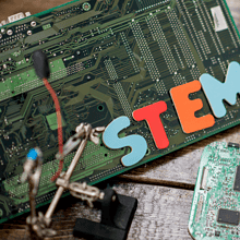 A variety of science-related materials are displayed on a table, including: a large motherboard, science flasks filled with color liquids, and various computer parts. “STEM” is spelling out in colorful letters.