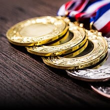 A close-up of several gold, silver, and bronze medals with red, white, and blue ribbons laid out on a wooden surface. These medals have intricate designs etched around the edges.