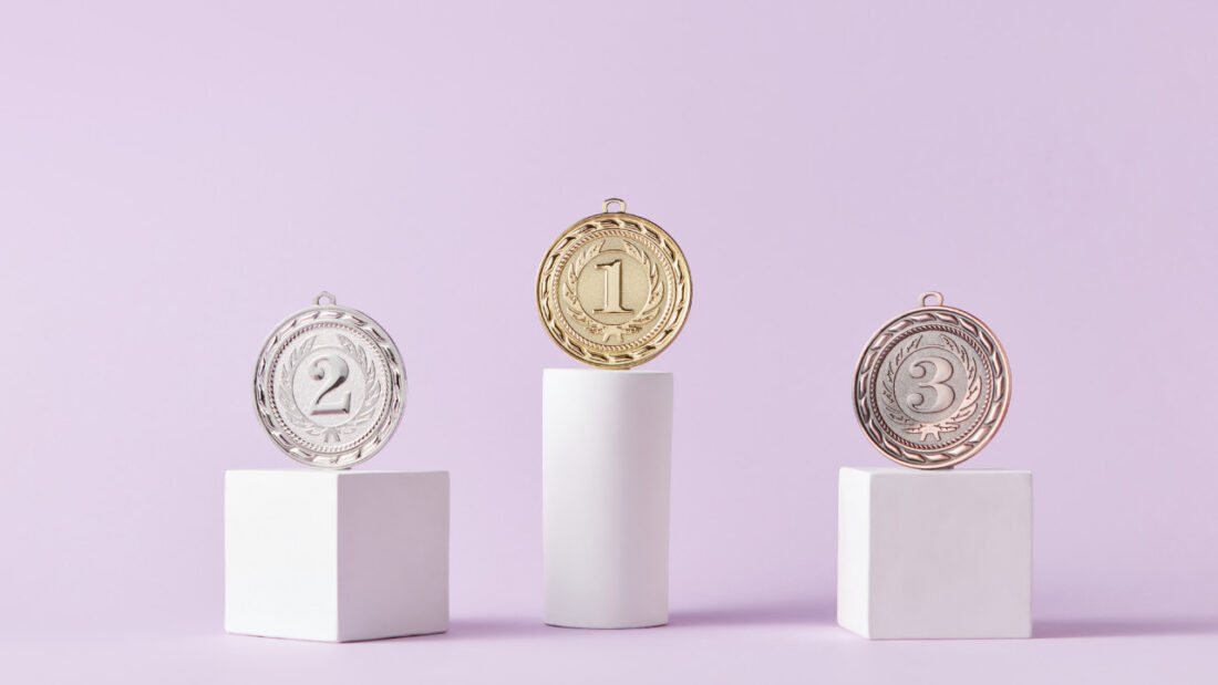 Three medals are positioned on white podiums against a light purple background. The gold medal with the number 1 is on the highest podium, the silver medal with the number 2 is on the left podium, and the bronze medal with the number 3 is on the right podium.