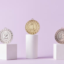 Three medals are positioned on white podiums against a light purple background. The gold medal with the number 1 is on the highest podium, the silver medal with the number 2 is on the left podium, and the bronze medal with the number 3 is on the right podium.