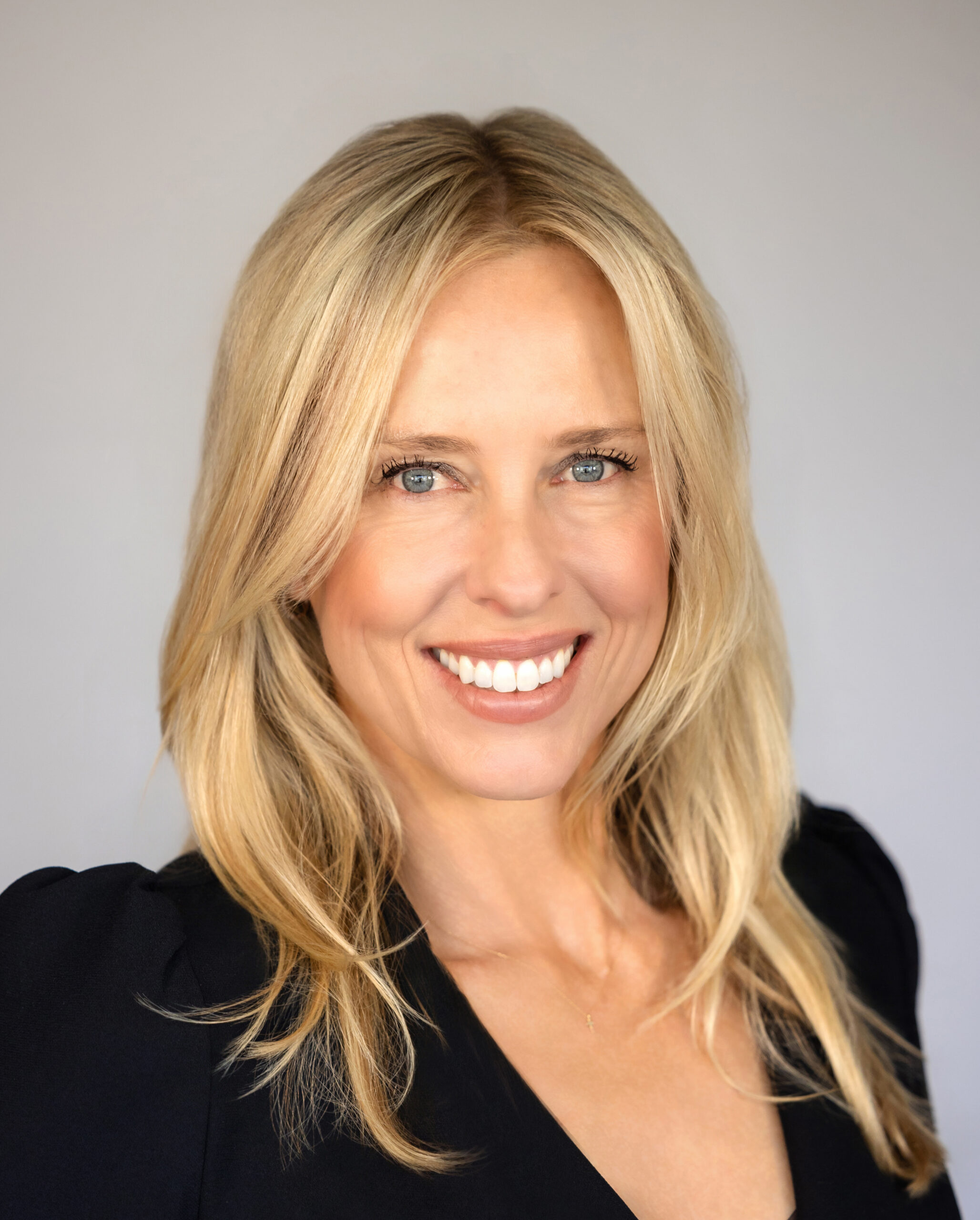 Smiling woman with long blonde hair and blue eyes, Heather Raney wears a black top as she poses in front of a plain, light gray background.